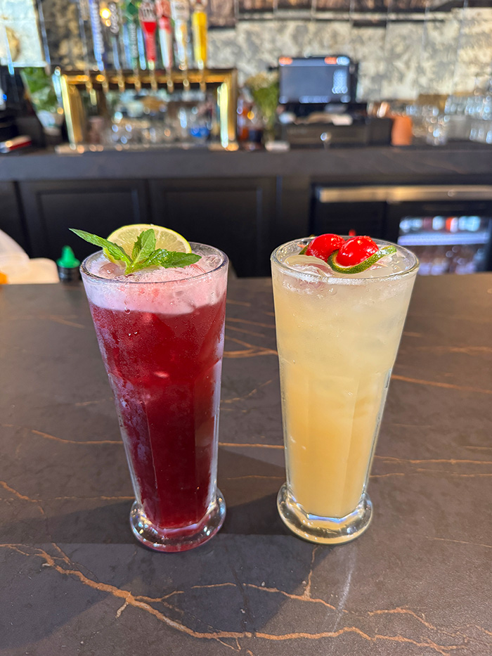 Two tall aguas frescas, jamaica and pineapple, side by side on a dark bar