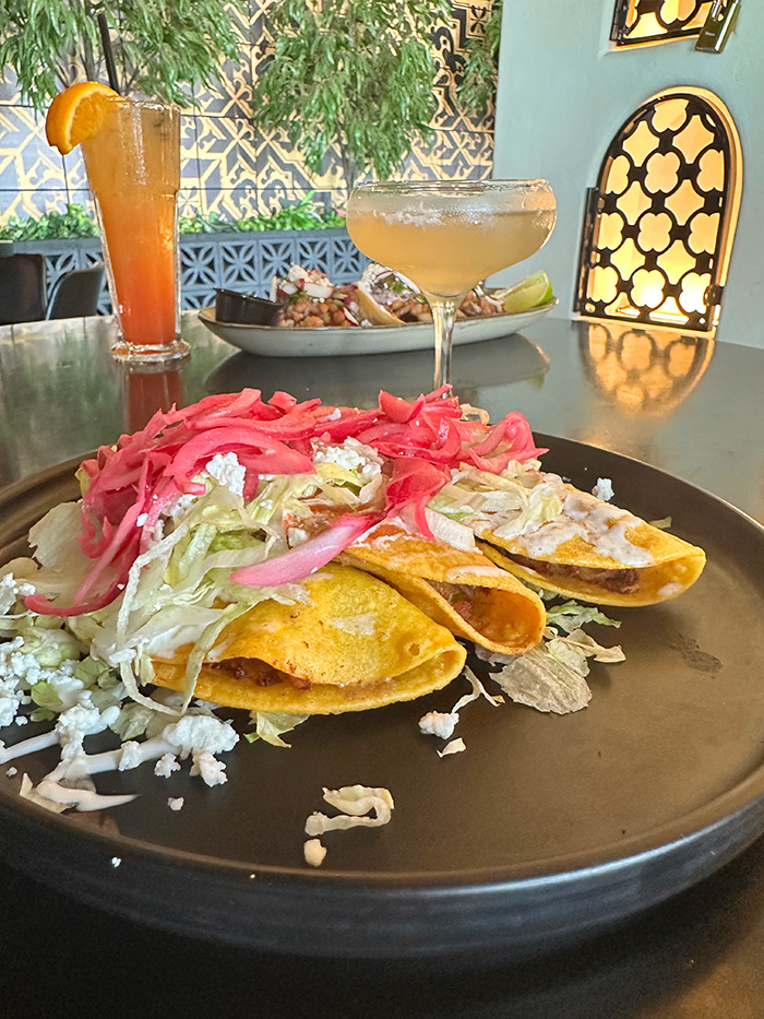 Two golden crispy tacos with pickled onions and queso fresco against a tile wall patio
