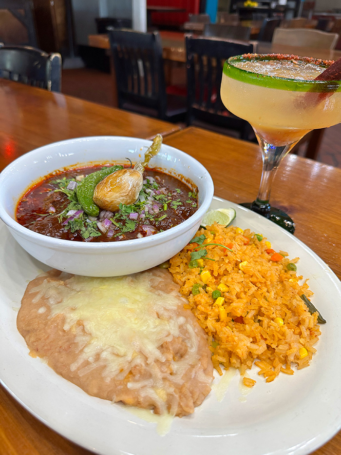 Bowl of birria consomé with garlic and jalapeño beside refried beans, rice and a margarita