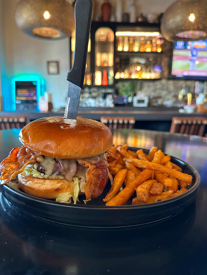 Qué Pasa Spicy Burger — half-pound Wagyu with bacon and pepper jack, knife standing in the brioche bun, sweet potato fries