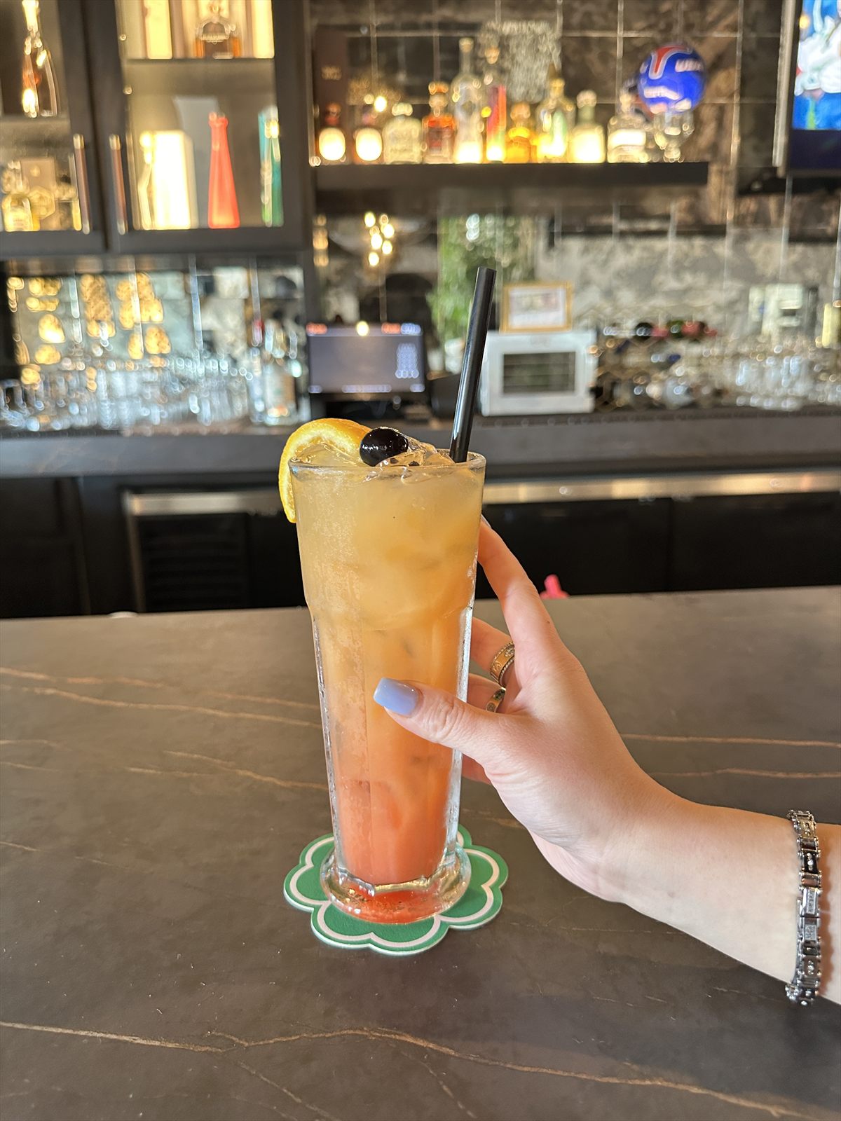 Tall tequila sunrise with orange wheel and cherry, held at a dimly lit bar